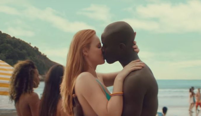 Mulher branca e homem negro se beijando em frente a uma praia
